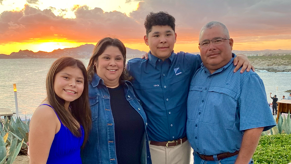 Jeffrey Torres, wearing a navy blue suit, stands smiling beside his family against a bright background; the group is dressed in semi-formal attire and appears proud and joyful, highlighting their connection and unity.